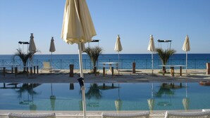 Ein Poolbereich mit Liegestühlen und Sonnenschirmen, mit Blick auf das Meer und einem ruhigen Pool, der die Umgebung spiegelt.
