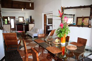 Ein geräumiger Wohnbereich mit einem Glastisch, umgeben von Holzsesseln, einem Sofa und einem Couchtisch. Im Hintergrund sind ein großer Spiegel und ein Topfpflanze zu sehen.