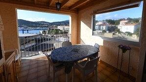 A balcony with a table and chairs, a hanging light fixture, and a view of a coastal town and hills. - Duplex, near the city center, on the left port exit (Port-Vendres)