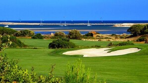 Ein Golfplatz mit einem sandigen Bunker, üppigem Grün und Blick auf das Meer mit Booten im Hintergrund.