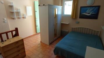 A bedroom with a bed, a wardrobe, a dresser, a radiator, a window with curtains, and a wall-mounted shelf.