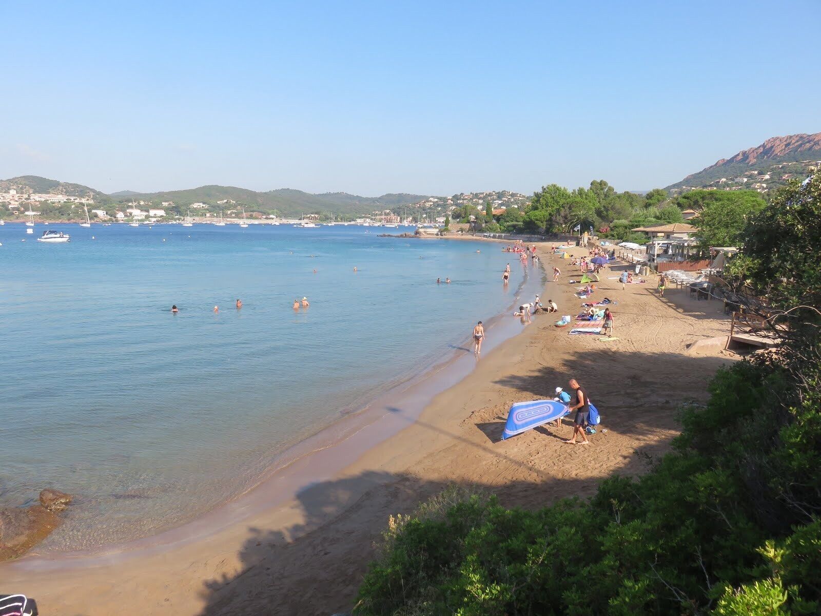 Une plage avec une eau d’un bleu limpide, des gens qui se baignent et une rive sablonneuse parsemée de parasols et de chaises de plage.
