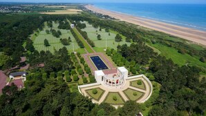 An aerial view of a landscaped estate with a central circular building, surrounded by meticulously maintained gardens and a beach in the background.