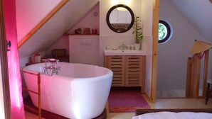A bedroom with a large white bathtub, a wooden wardrobe, a small bathroom with a sink and mirror, and a window with a view of greenery.