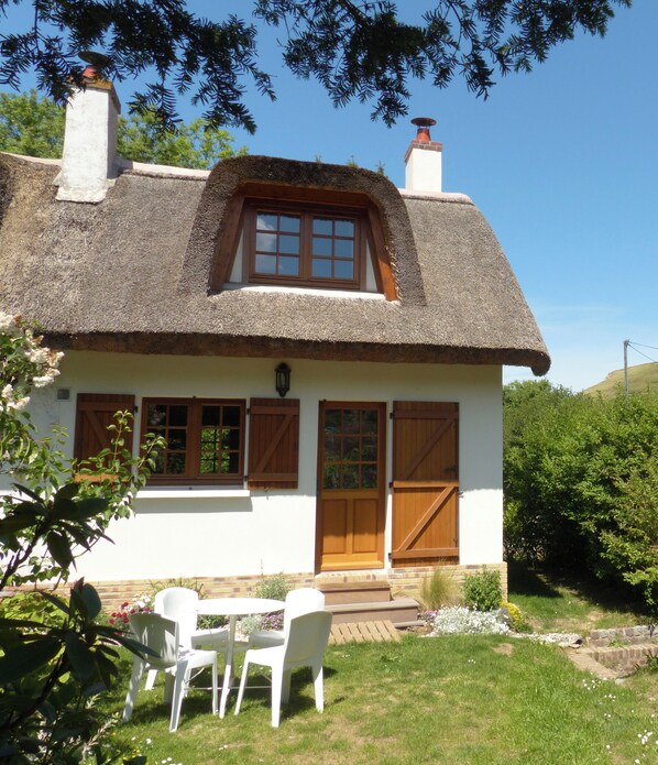 Een traditioneel huis met rieten dak, een houten deur en ramen, omringd door groen en een heldere lucht.