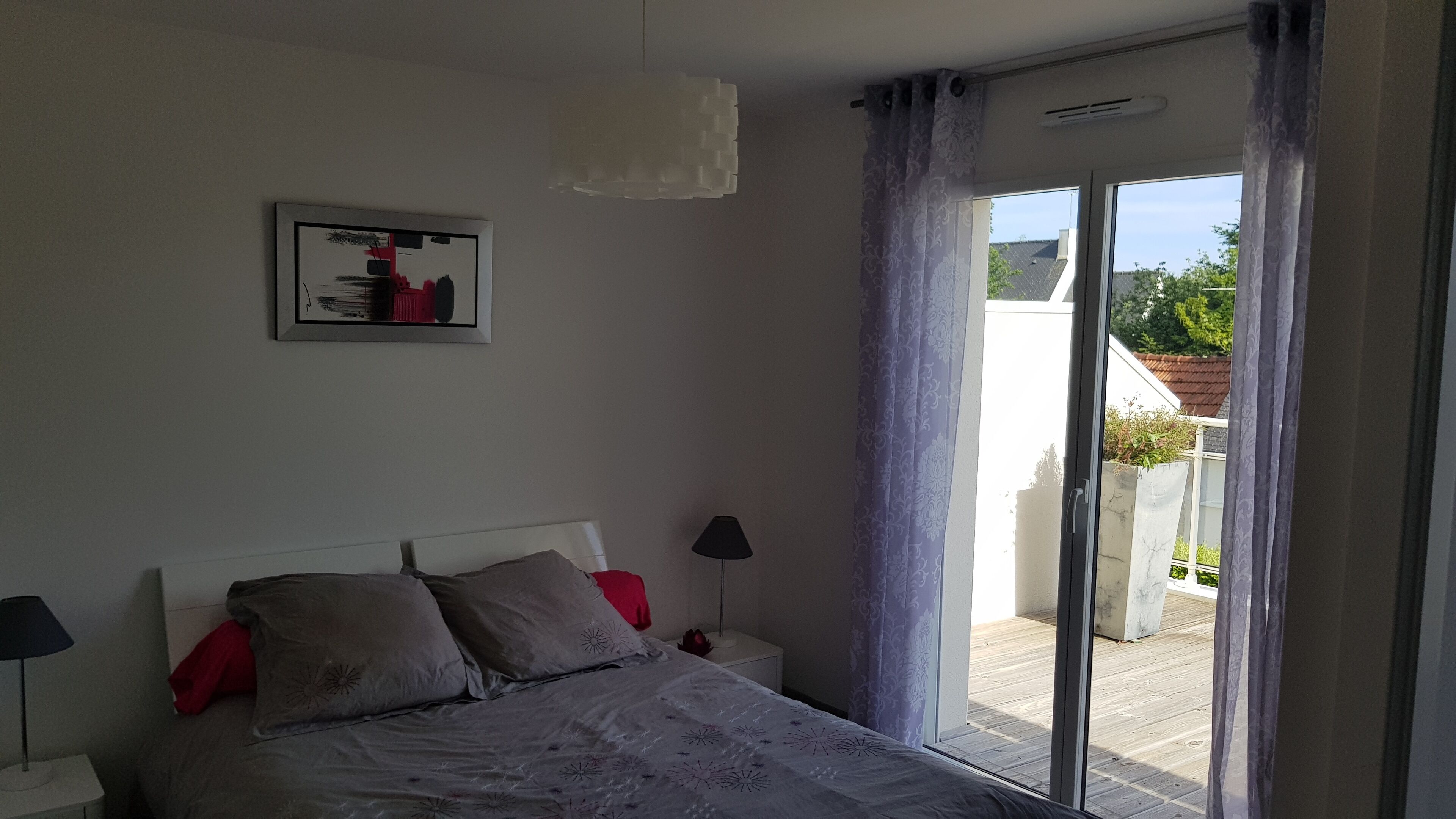 A bedroom with a bed, a nightstand, a lamp, a window with curtains, and a view of the outside.