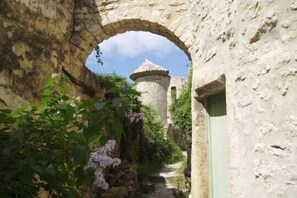 A narrow stone pathway leading to a tower-like structure with a conical roof, surrounded by greenery and a stone wall.