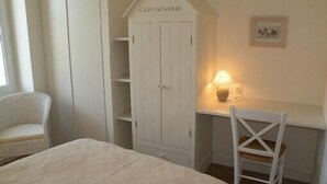 A bedroom with a bed, a white wardrobe labeled "CAPITAINEERIE," a desk with a lamp, and a white chair.