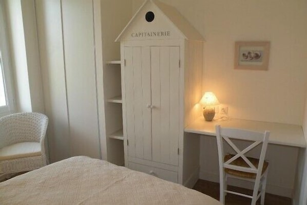A bedroom with a bed, a white wardrobe labeled "CAPITAINEERIE," a desk with a lamp, and a white chair.