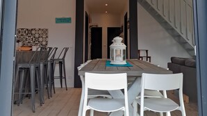 Een eethoek met een houten tafel en witte stoelen, een trap en een keuken met betegelde vloer.
