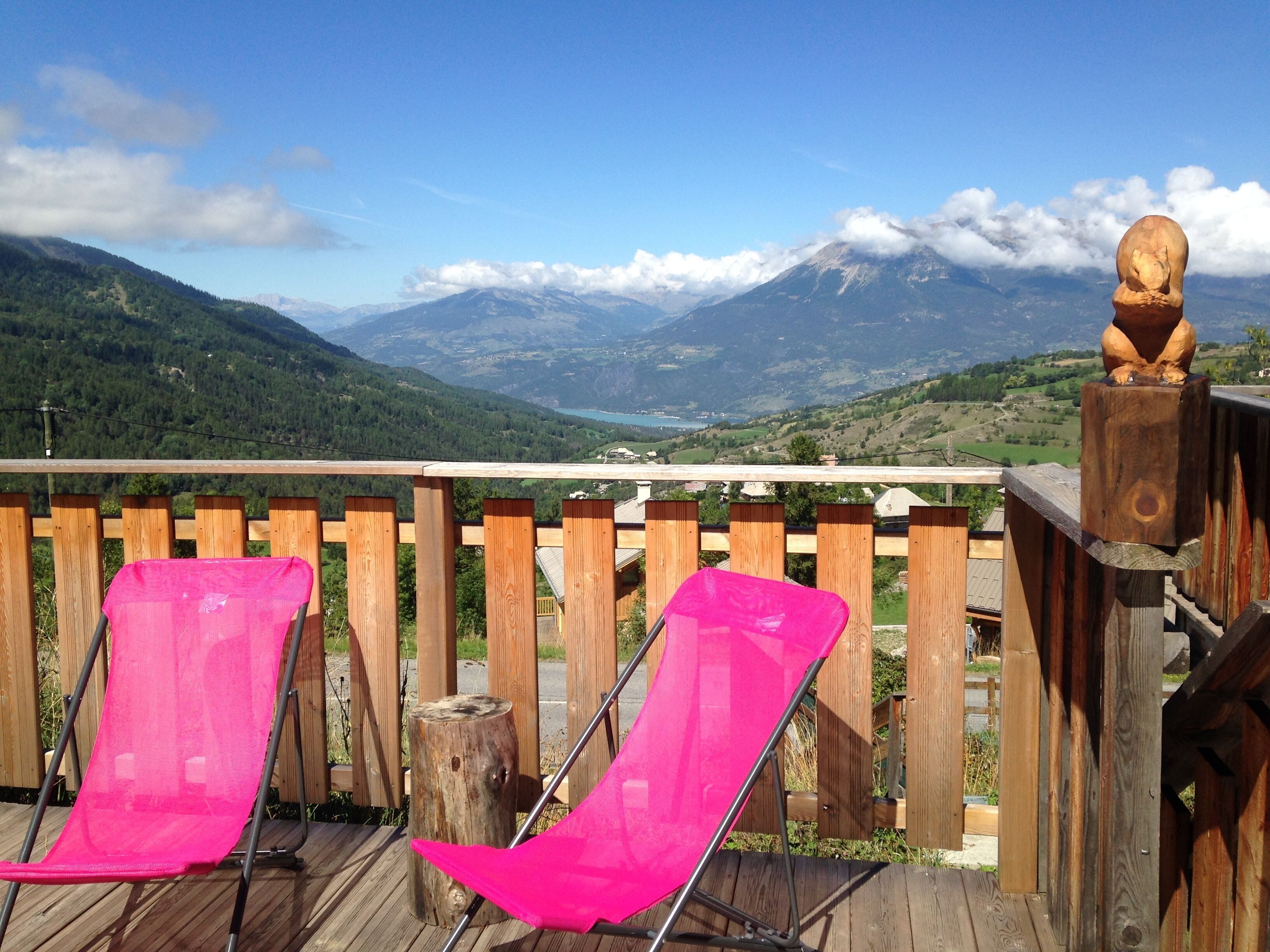 A wooden deck with two pink chairs, a wooden fence, and a view of mountains and a lake.