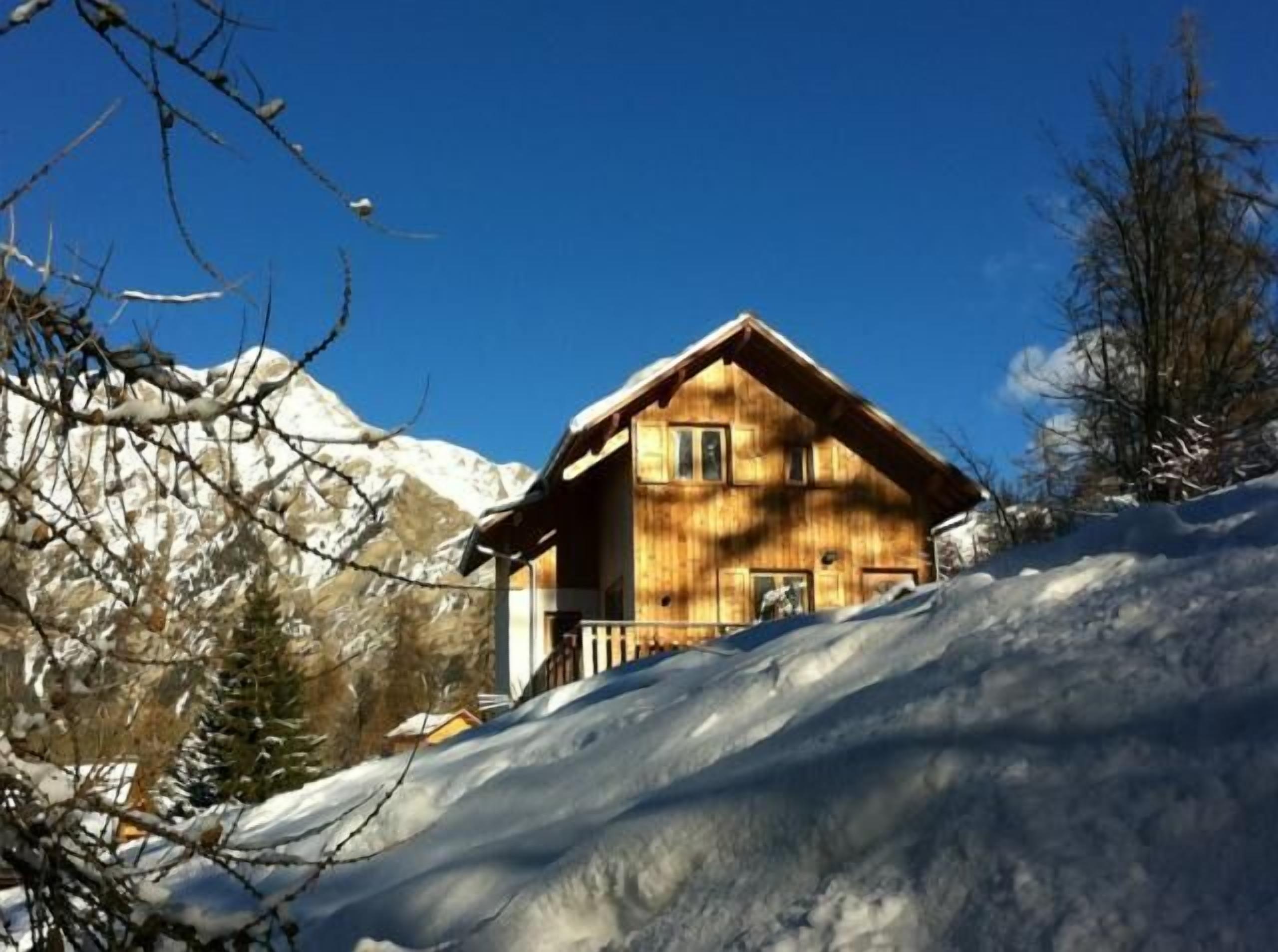 Eine Holzhütte mit geneigtem Dach, umgeben von schneebedeckten Bergen und Bäumen.