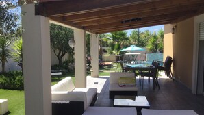 Een terras buiten met een pergola, lounge fauteuils, een tafel en een zwembad op de achtergrond.