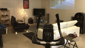 Een woonkamer met een loopband, een witte bank, een zwarte fauteuil, een televisie en een projectiescherm waarop een landschap wordt vertoond.