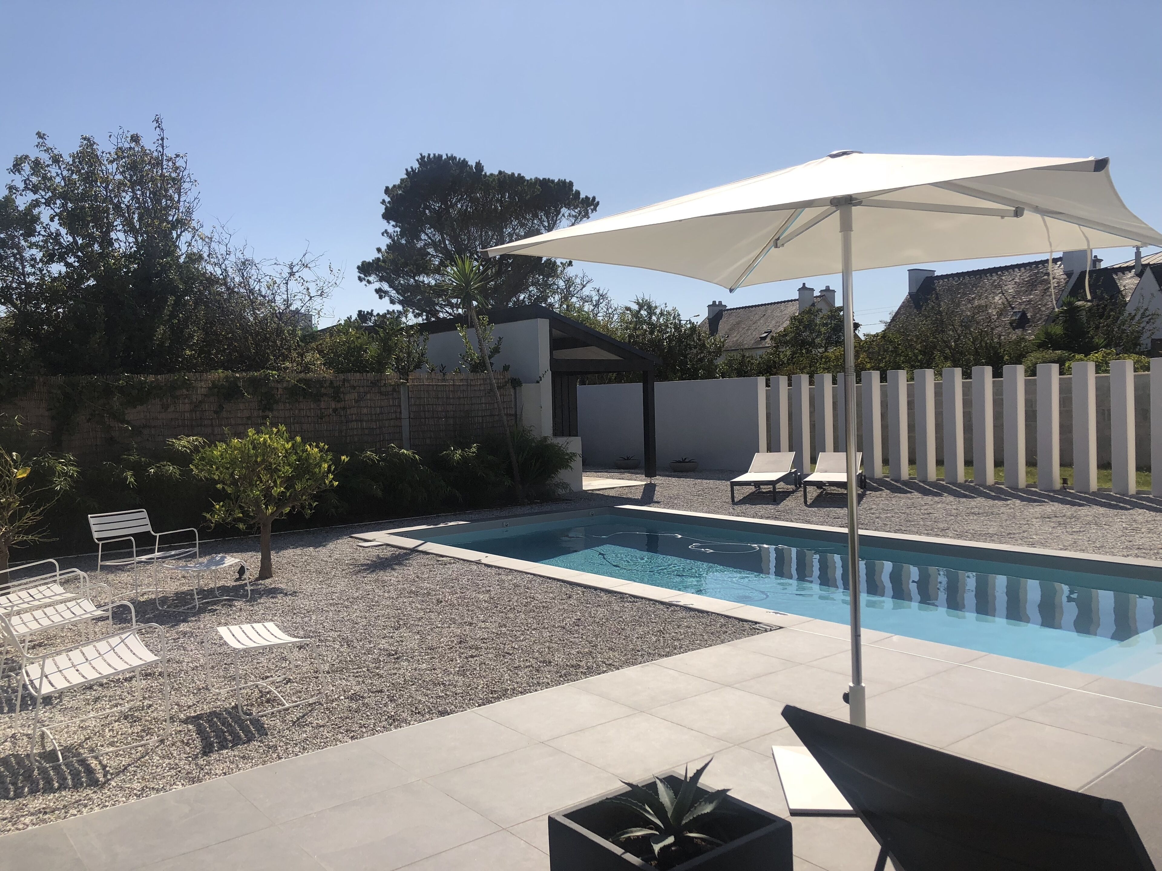 A swimming pool with a white umbrella, surrounded by lounge chairs and a small garden area with gravel and plants.