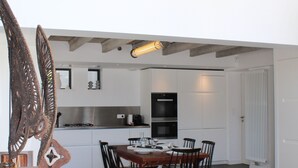 Ein Essbereich mit einem Holztisch und -stühlen, einem großen Fenster darüber und einer Küche im Hintergrund.