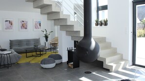 Ein modernes Wohnzimmer mit einem grauen Sofa, einem Couchtisch und einem schwarzen Kamin. Eine Treppe führt in ein Obergeschoss, und ein Fenster ist mit Pflanzen dekoriert.