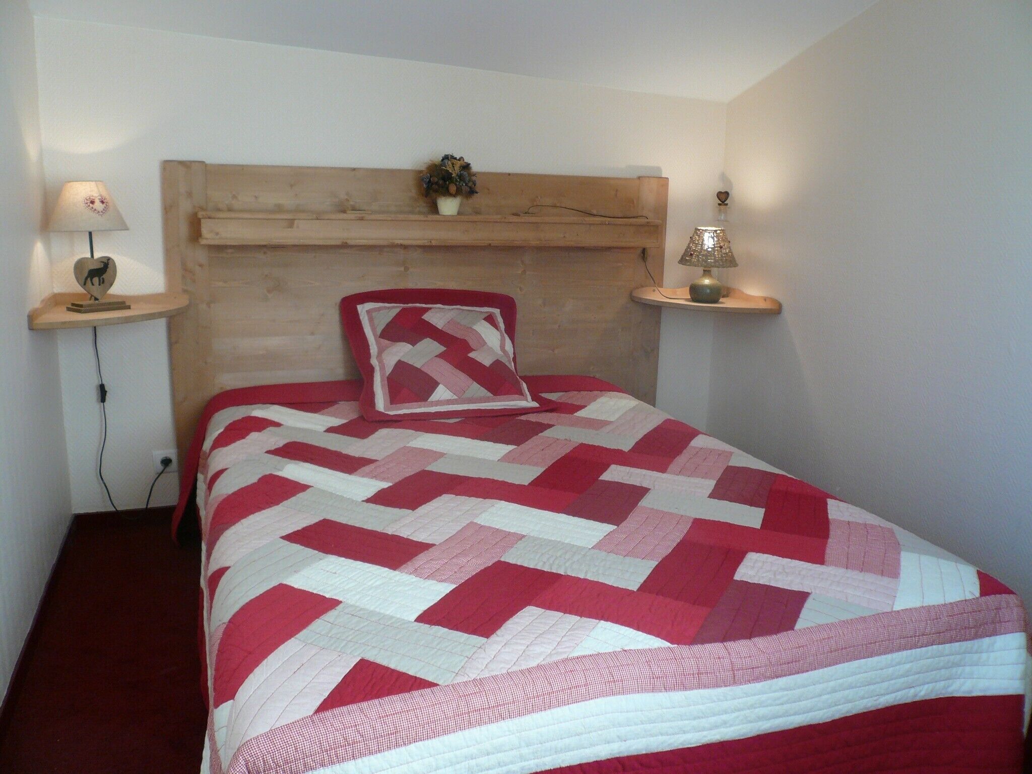 A bedroom with a wooden headboard, a red and white quilt, a heart-shaped pillow, bedside lamps, and a small plant on the nightstand.