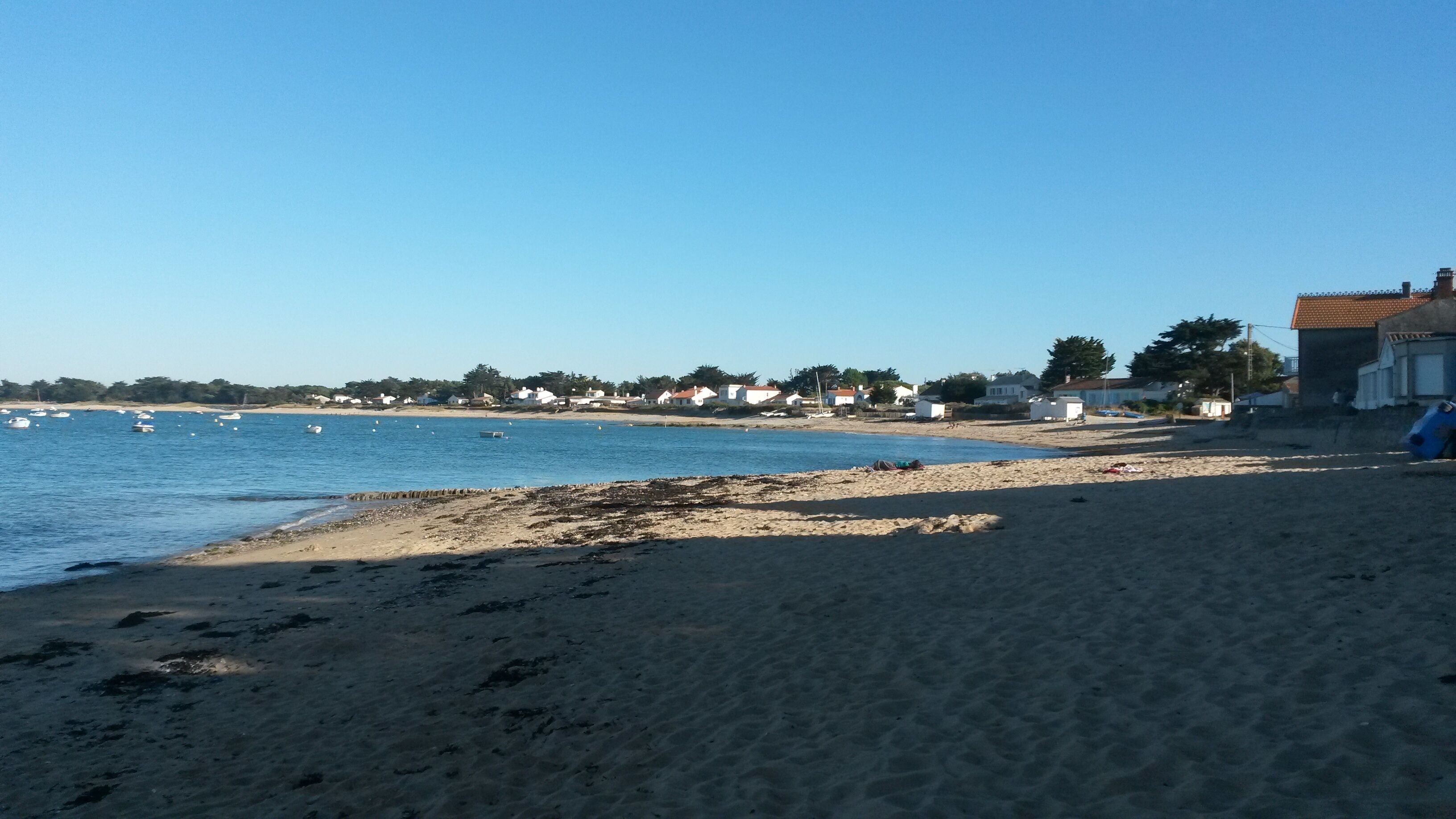 A sandy beach with a clear blue sea, a few boats, and houses in the background.