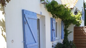 Ein weißes Gebäude mit blauen Fensterläden, einer Holzterrasse und einem kleinen Garten mit einer Weinrebe und Blumen.