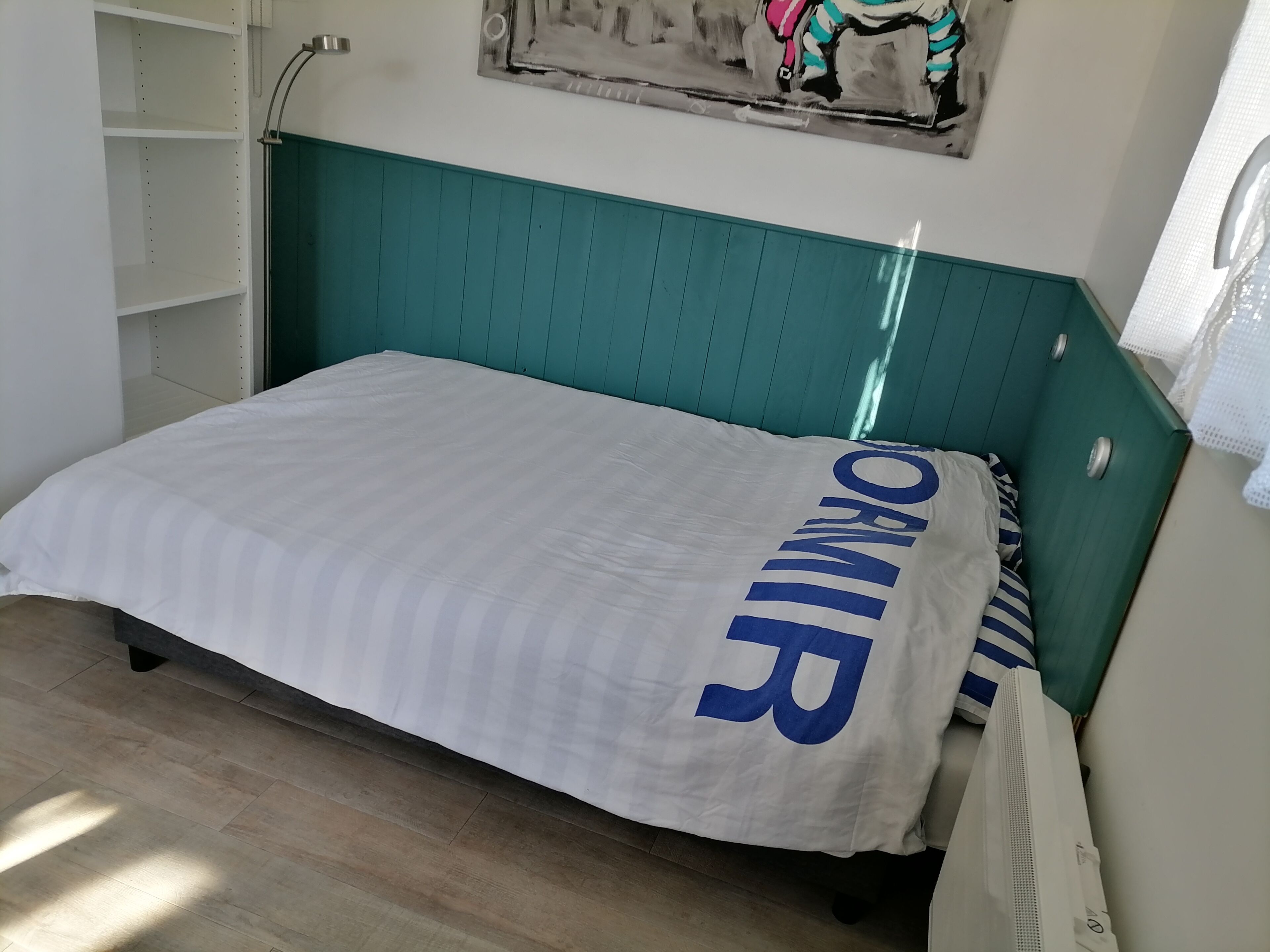 Una cama con colcha blanca y azul, cabecera color turquesa y una estantería fijada a la pared.