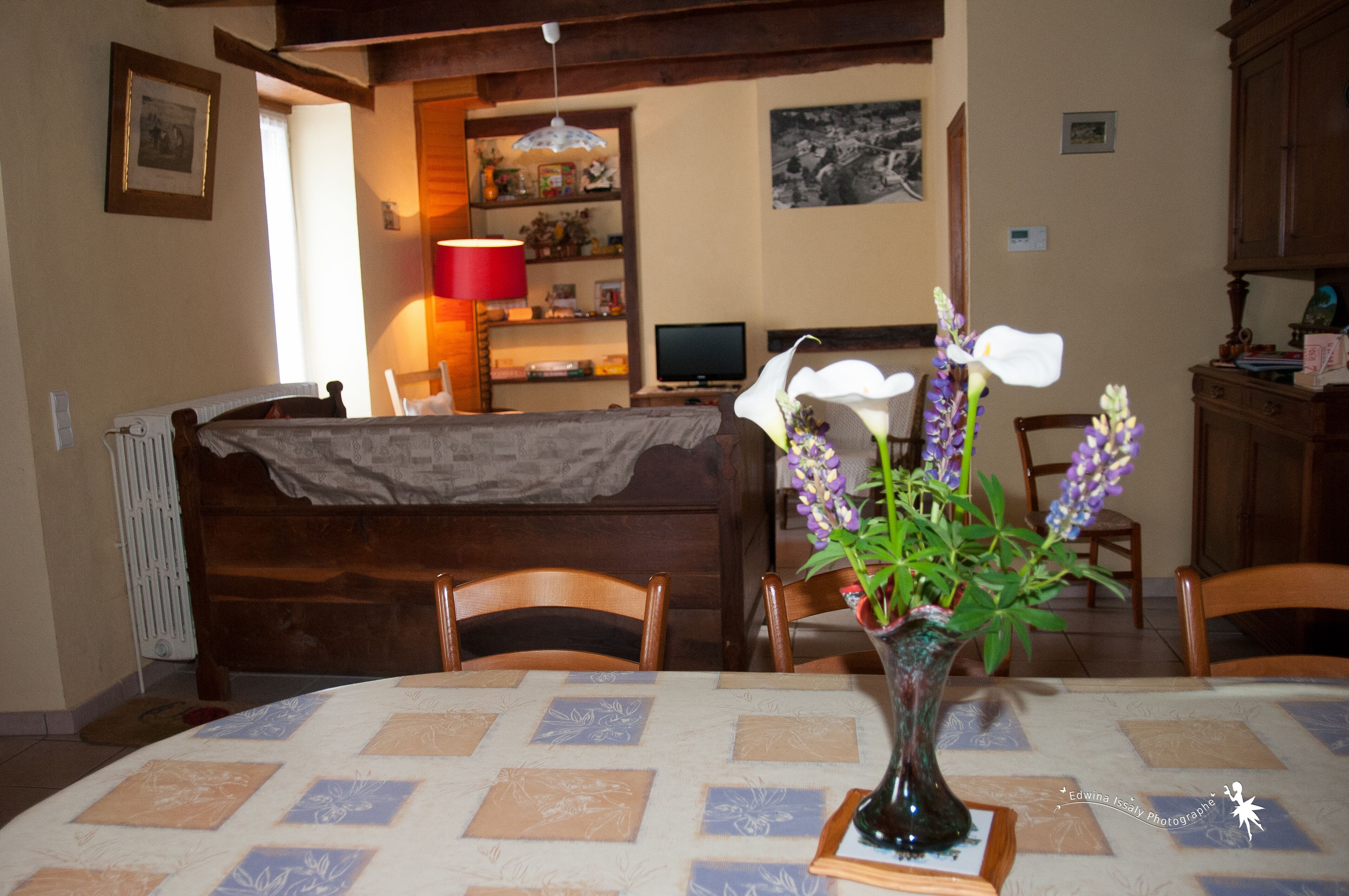 Un coin repas avec une table dressée pour deux personnes, un vase rempli de fleurs et un téléviseur en arrière-plan.