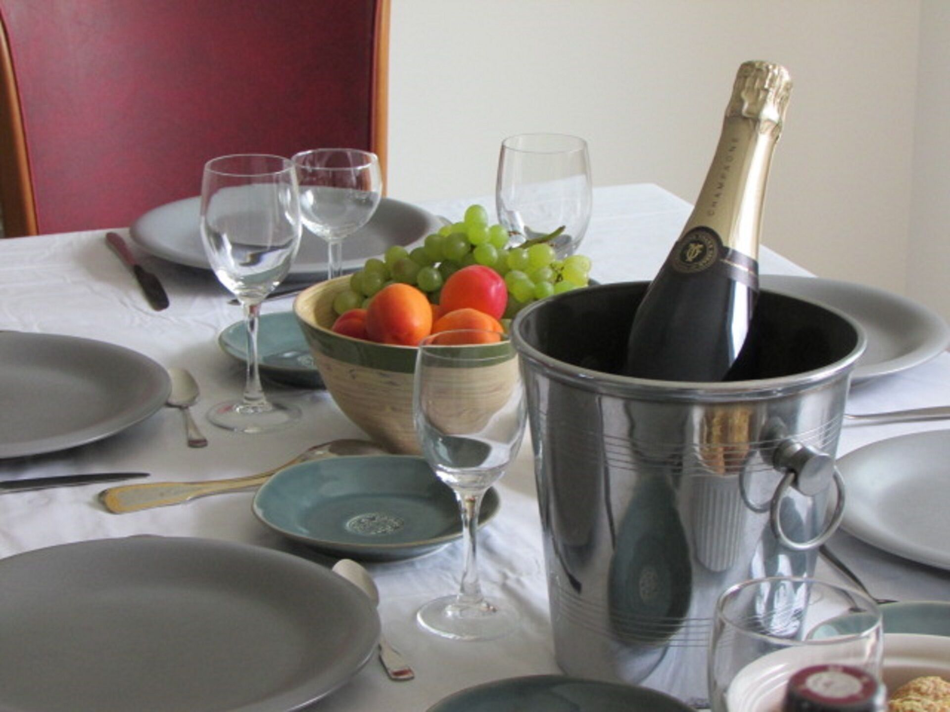 Ein Esstisch ist gedeckt mit Tellern, Gläsern, einer Obstschale und einer Champagnerflasche in einem Kübel.