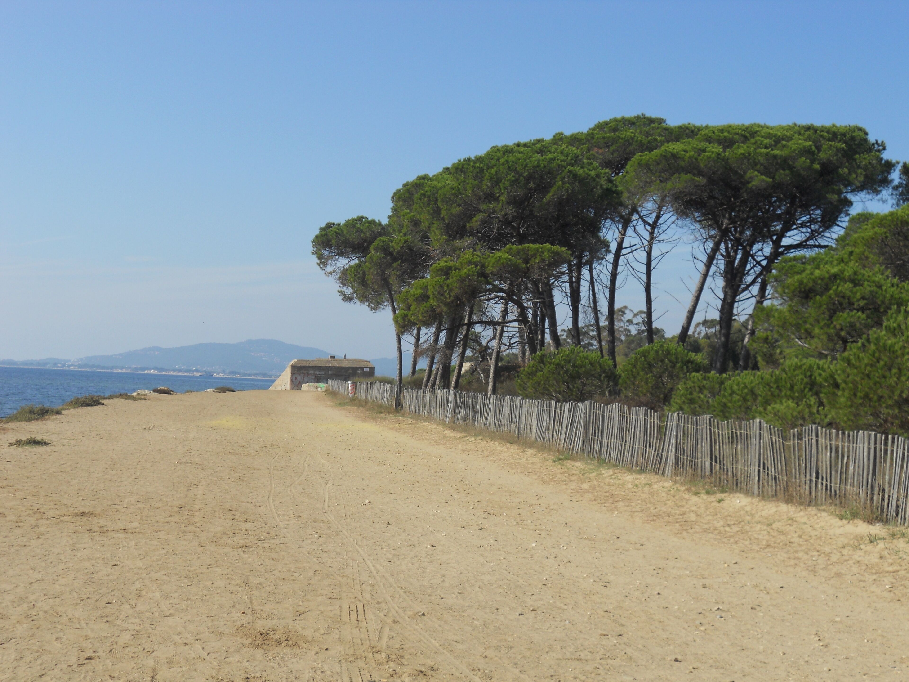Un sendero de arena con una cerca de madera y un pequeño edificio, probablemente una cabaña de playa, con una vista despejada del mar y montañas distantes.