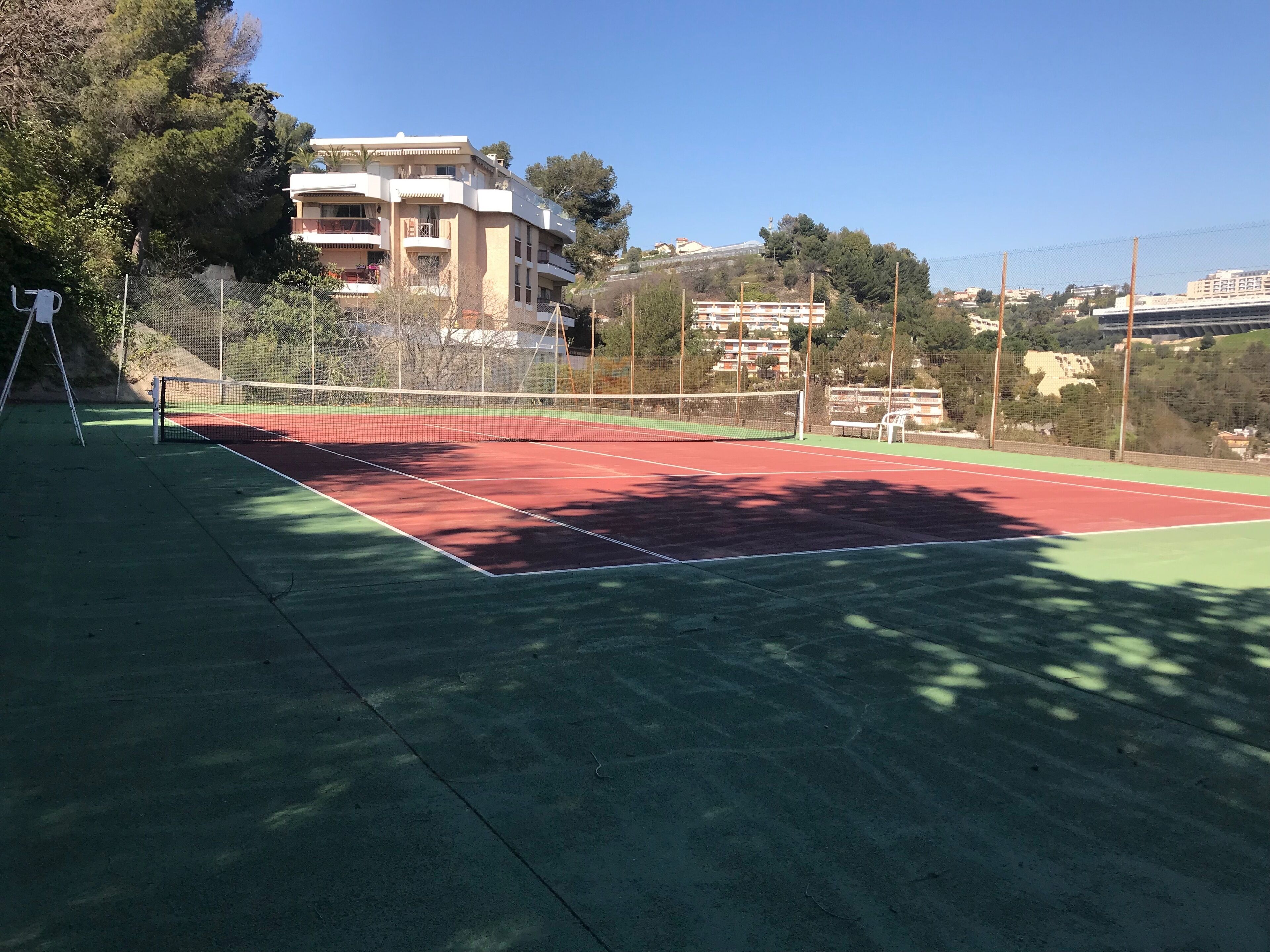 Een tennisbaan met een net, omheind door een hek, en gebouwen op de achtergrond.