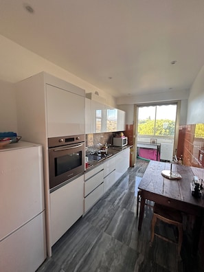 A modern kitchen with white cabinets, a built-in oven, and a gas stove. There is a dining table with chairs and a view of greenery outside. - ✨ Comfort & sun in Nice 🌞 | Terrace sea view, pools & tennis (Nice)