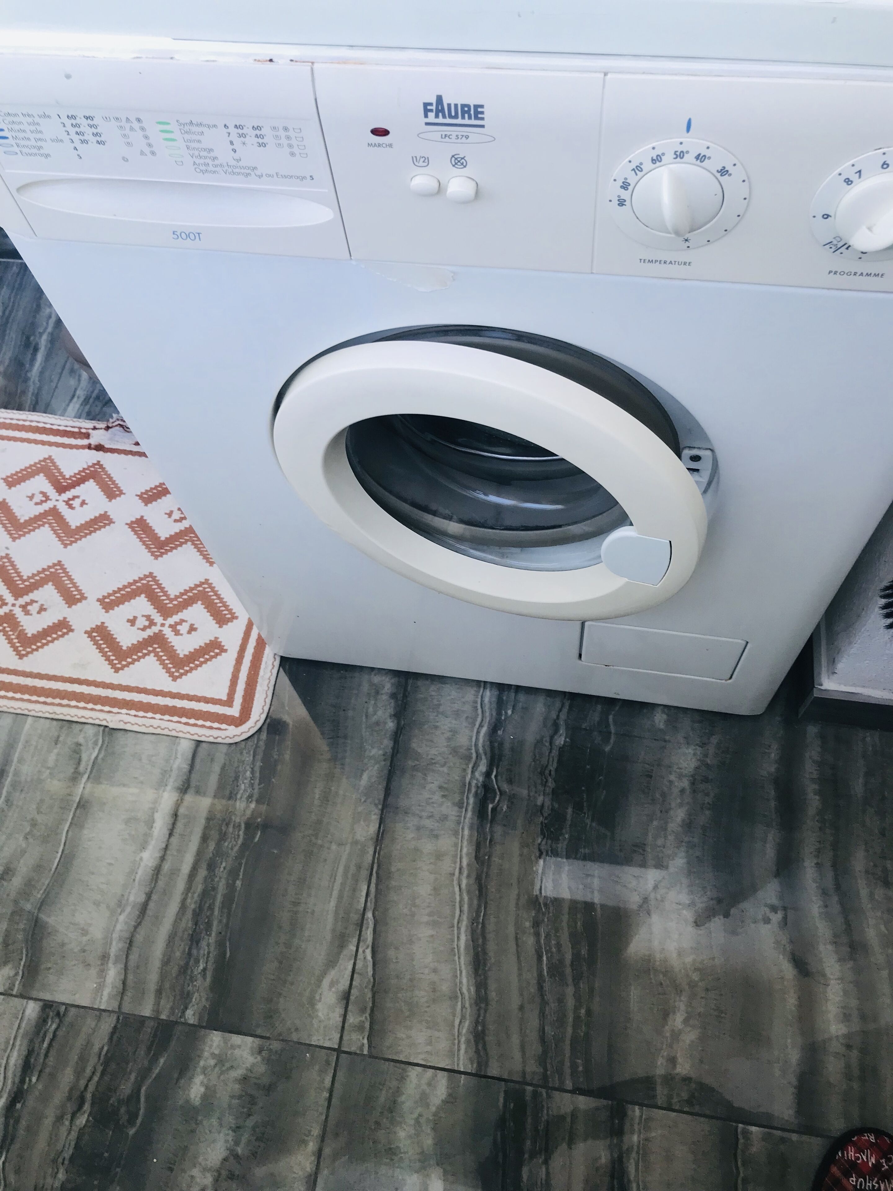 Vue de face d’un lave-linge Faure blanc, doté d’un panneau de commande, posé sur un sol carrelé et surmonté d’un tapis à motifs.