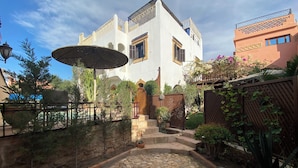 Un patio con un camino de piedra, una valla de madera y un edificio con fachada blanca y contraventanas azules.