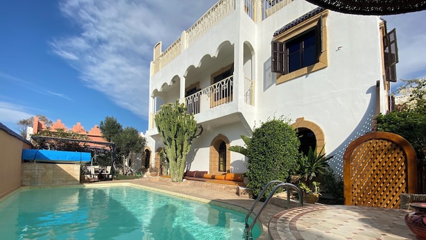 Un área de piscina con una piscina azul cristalina, un espacio de descanso cubierto y un edificio con ventanas arqueadas y balcones.