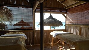 Una habitación con dos mesas de masaje, techo de paja y vistas a la playa.