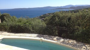 A swimming pool with a stone deck, surrounded by lush greenery and overlooking a coastal landscape with hills and the sea. - VILLA WITH SEA VIEW ON THE BAY OF ST TROPEZ (Roquebrune-Sur-Argens)