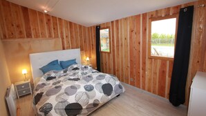 A bedroom with a wooden wall, a bed with a patterned bedspread, a nightstand with a lamp, and a window with curtains.