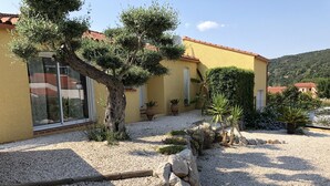 A yellow building with a red-tiled roof, surrounded by gravel, trees, and potted plants. - Very beautiful wooded villa, heated swimming pool, breathtaking view - 8/10 people (Les Cluses)