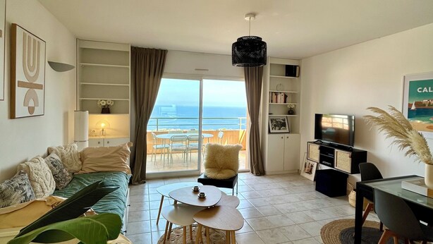 Un salon spacieux doté d’une grande fenêtre offrant une vue sur la mer, un coin salon confortable avec un canapé et une petite table, ainsi qu’un téléviseur posé sur un meuble.
