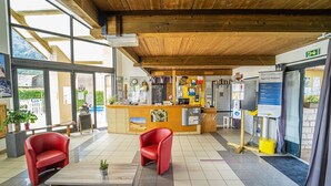 A reception area with a wooden reception desk, red chairs, a small table, and a large window. - Logement 4/6 pers. - Blajoux (Quézac)