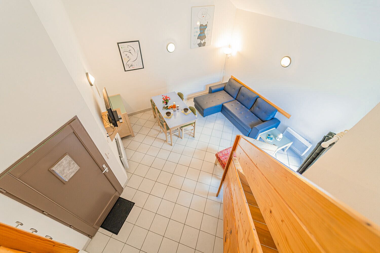 A high-ceilinged living room with a blue sofa, a dining table with chairs, and a staircase leading to an upper floor.
