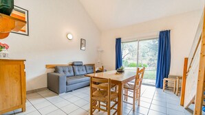 A dining area with a wooden table and chairs, a grey sofa, and a view of the outdoors through large windows with blue curtains. - Logement 4/6 pers. - Blajoux (Quézac)