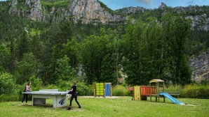 An outdoor area with a ping pong table, a playground with a slide, and a mountain backdrop. - Logement 4/6 pers. - Blajoux (Quézac)
