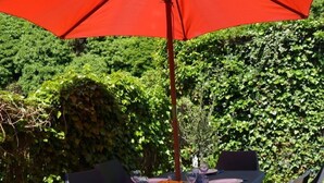 Un espace de restauration en plein air avec une table et des chaises, un parasol rouge et un jardin luxuriant en arrière-plan.