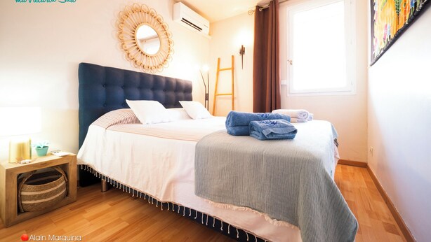 Un dormitorio con cabecera capitonada, un espejo redondo, una cama con ropa de cama blanca y a rayas, una mesita de noche de madera con una lámpara, y una ventana con cortinas.
