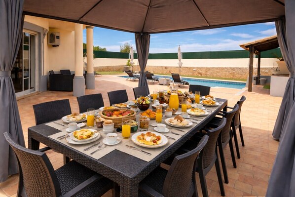 A covered outdoor dining area with a pool view, wicker chairs, and a table set for breakfast.