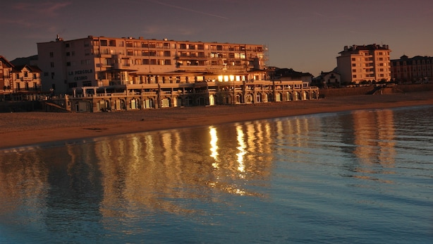A coastal hotel with multiple floors, a beach in the foreground, and a pier extending into the water.