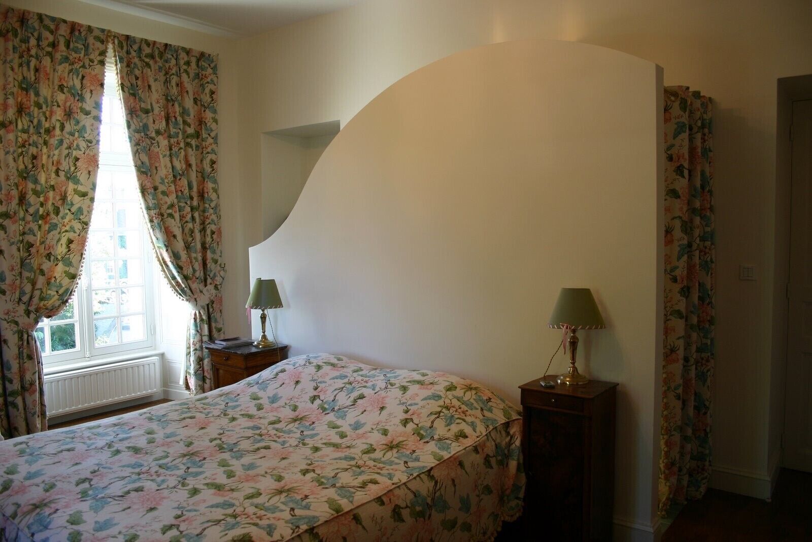 A bedroom with a bed, two bedside tables, floral curtains, and a window with a view.
