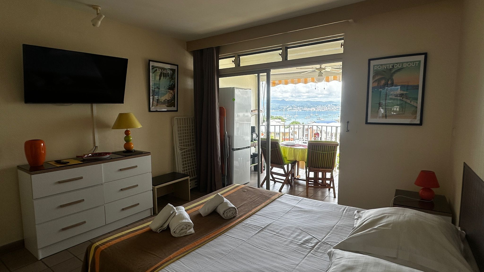 Ein Hotelzimmer mit Bett, Fernseher, Schreibtisch mit Lampe, Kommode, Balkon mit Tisch und Stühlen sowie Blick auf das Meer.