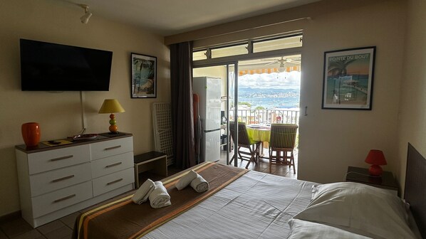 Habitación de hotel con cama, televisión, escritorio con lámpara, cómoda y balcón con mesa y sillas, y vista al mar.