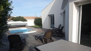 Une terrasse en bois avec une piscine, du mobilier d’extérieur et un bâtiment blanc doté d’une porte coulissante.